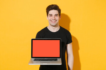 Smiling young man guy 20s in casual black t-shirt posing isolated on yellow background studio portrait. People lifestyle concept. Mock up copy space. Hold laptop pc computer with blank empty screen.