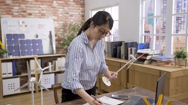 young asian chinese woman engineer holding and checking wind turbine models in office standing at work desk. elegant female architect examining windmill miniature and write on notebook with new ideas