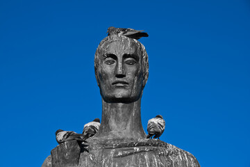 Monumento em frente à Catedral de Brasília. Estátua com pássaros na cabeça e céu azul ao fundo.