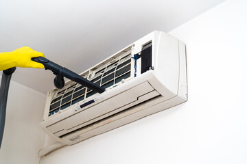 A worker is using a vacuum cleaner to clean the air conditioner. A white air conditioner is installed on a white wall in the apartment