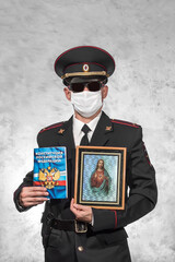 Russian police officer in uniform in a white medical mask and black glasses and holds Jesus Christ...