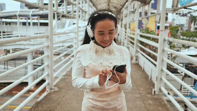 A cheerful Indonesian girl is standing on a bridge in the city in white headphones and choosing music on her smartphone.