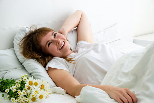 Happy young woman with bouquet of daisies is lying in bed at home