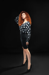 Isolated slender red-haired curly girl in a blouse and a black skirt against a black background. Office employee.