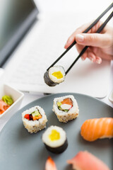 Asian sushi set with laptop and female hand holding chopsticks. Healthy meal at work food delivery concept.