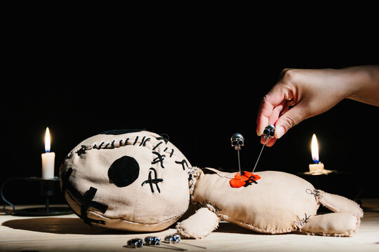 Woman's hand stabbing with a pin a voodoo doll during occult magic ritual on a black background with candles