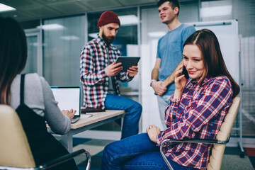 Successful smiling hipster girl making positive telephone call via application on cellular with two guys on back view in break of  studying with male and female students at modern class room.