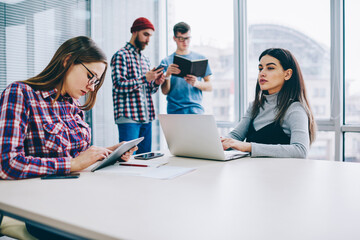 Serious hipster girl thoughtful looking away in break of working on laptop computer while her female friend in spectacles typing text message using digital tablet with wifi connection sitting desktop.