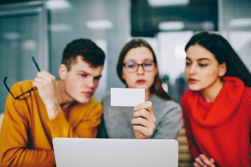Pensive young people dressed in casual wear reading information on blank card sitting in office at netbook.Selective focus on business card with mock up area for your advertisement in woman's hand