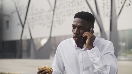 African American Businessman talking on cell phone and eating sandwich in park near office