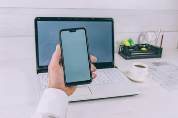 Mock-up Technology. Businessman holds a smartphone in his hands, on a background of laptop and coffee on a wooden table.