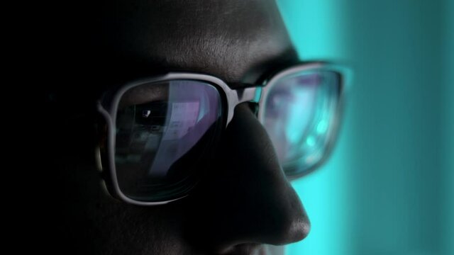 Male eyes in eyeglasses late at night scrolling in front of laptop. Coder, programmer or developer using laptop in dark. Close up of glasses with reflection of computer screen.