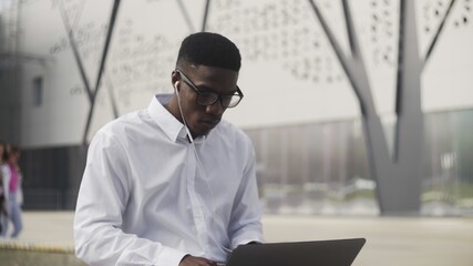 African Businessman with headphones Sitting on bench in park and working on notebook