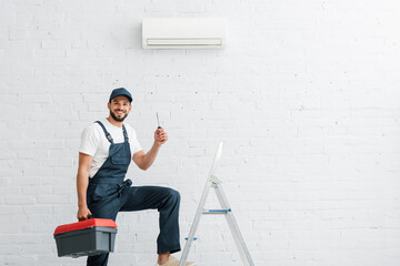 Smiling workman in uniform holding screwdriver near ladder and air conditioner on wall