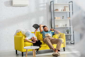 Selective focus of exhausted couple with fan and book feeling hot in living room