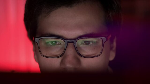 Man in eyeglasses late at night scrolling in front of laptop profile shot on red background. Coder, programmer or developer using laptop in dark.
