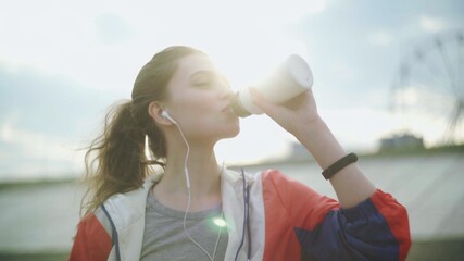 Brunette teenager girl drinking cold water after running in the park. Beautiful fitness athlete...