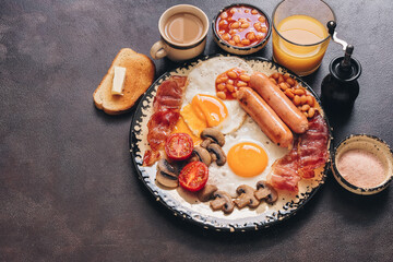 English traditional breakfast on a dark brown rustic background, copy space. Fried eggs with bacon, sausages and beans, coffee, orange juice.