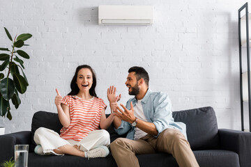 Selective focus of smiling woman showing thumbs up near cheerful boyfriend in living room
