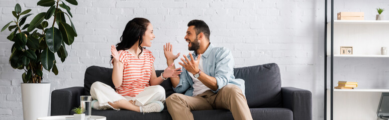 Panoramic crop of cheerful couple looking at each other on couch at home