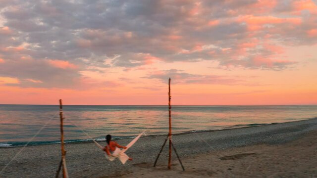 Bellissima ragazza va in vacanza sull'altalena in riva al mare calmo con nuvole e cielo arancione e rosso.