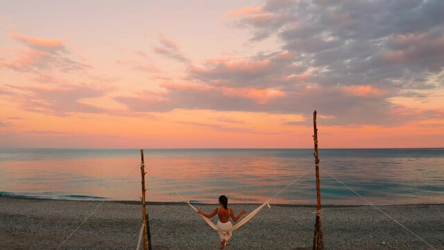 Bellissima ragazza va in vacanza sull'altalena in riva al mare calmo con nuvole e cielo arancione e rosso.