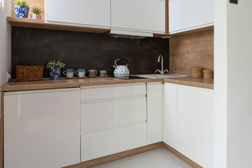 Small white kitchen with bright furniture and dark marble, wooden counter, plant and kettle. Simple interior of kitchen.