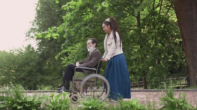 Adorable satisfied helpful young girl with dreadlocks pushing wheelchair with respected bearded elderly disables-man,walking in beautiful urban park
