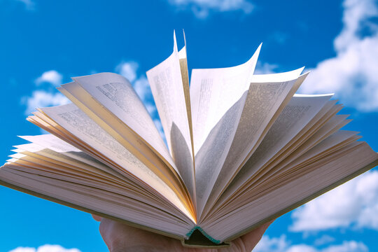 An open book in a hand raised to the sky. Blue sky with clouds create a background for the book.