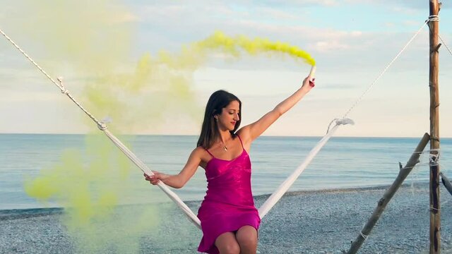 Ragazza con fumogeno giallo e vestito fucsia si dondola nell'altalena in riva al mare
