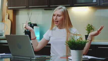 Cheerful woman singing and gesturing while looking on the computer screen. She spending her free time in the kitchen.