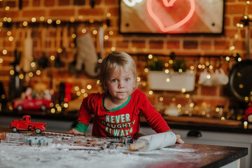 Fototapeta na wymiar Portrait of little blond boy in red Christmas pyjamas making cookies at family kitchen against Christmas lights.