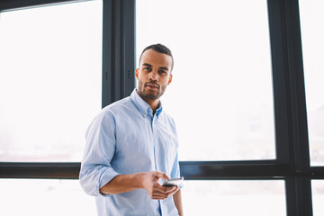 Fototapeta premium Half length portrait of dark skinned businessman in casual wear looking at camera while standing in loft office, handsome prosperous afro american entrepreneur checking for message holding smartphone