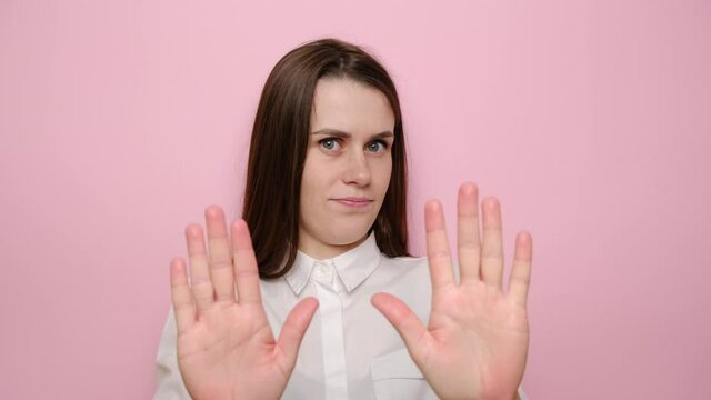 Portrait of young woman looking and points at camera waving hands do not wanna listen showing stop gesture, dressed in shirt, isolated on pink studio background. People emotions lifestyle concept