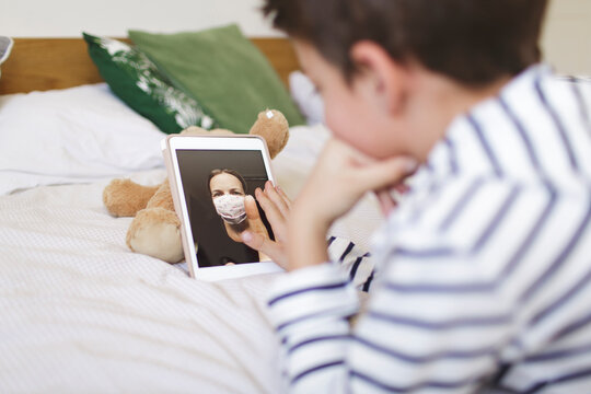 Boy lying on bed having video call on digital tablet with his mother wearng protective mask