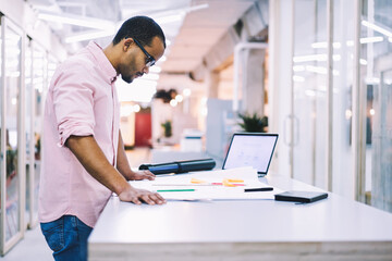 Mindful male engineer making correction in blueprint preparing for rebuilding using architectural application on laptop computer with mockup screen connected to fast 5G wireless in coworking office