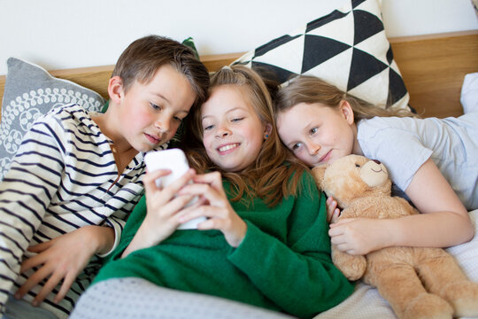Group picture of three children lying together on bed looking at cell phone