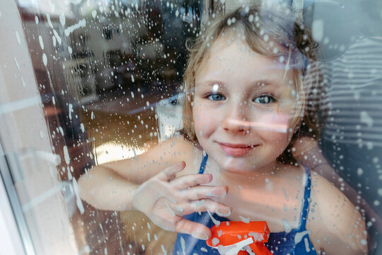 Portrait of smiling blond girl with spray bottle looking through wet window