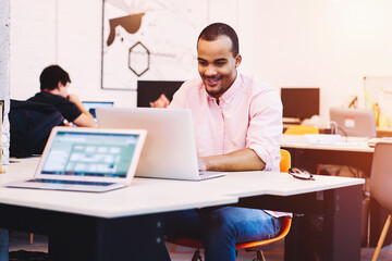 Cheerful afro american hipster guy checking email and chatting with friends in coworking space on...