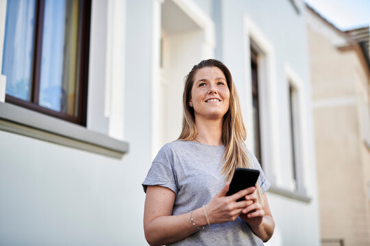 Woman looking away while holding mobile phone against house