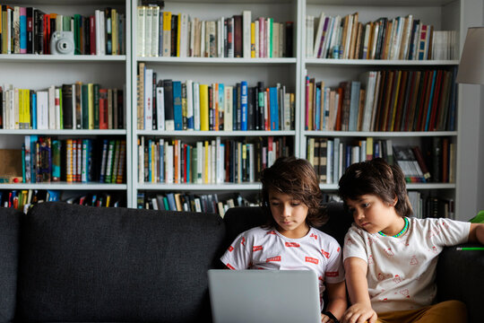 Brothers sitting with laptop on sofa against bookshelf in living room at home