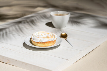 Lemon meringue little pie on a wooden board, table on a palm beach outdoor cafe in the shadows of palm branches. Continental breakfast with a cup of coffee.