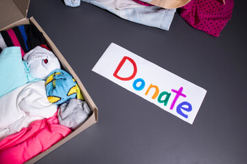 'Donate' sign handwritten with multicoloured letters. A box with clothes and a pile of clothes nearby on a grey table.clothes donation concept. copy space. High quality photo