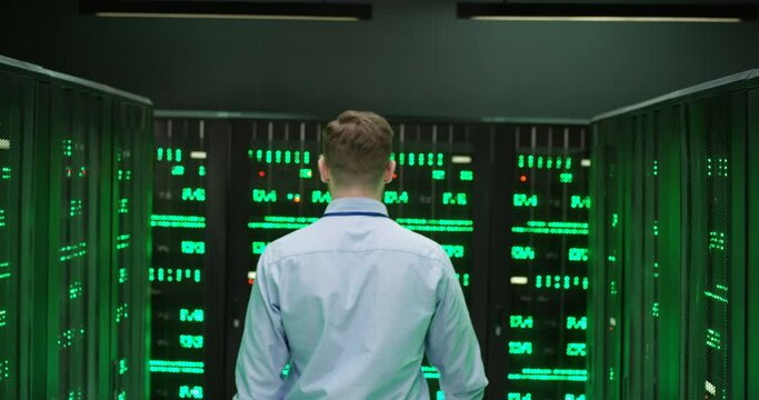 Rear of Caucasian young man walking among servers and checking work of big data processors. Back view. Male analytic in data storage strolling in information security office.
