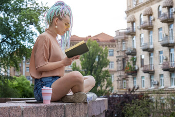 Stylish young woman with dreadlocks hairstyle and glasses reading a book while sitting against the backdrop of the city.