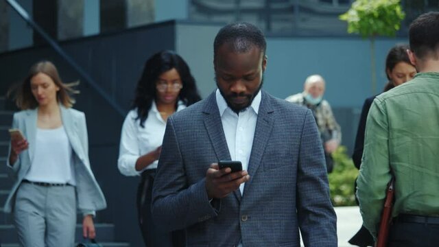 Handsome black young business leader entrepreneur using smartphone outdoors. Crowd of corporate busy co-workers with mobile phones having break. Business district.