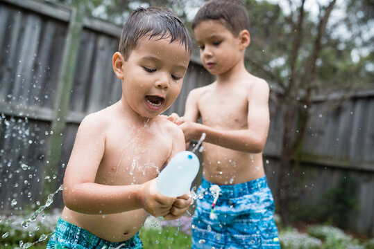 3 year old and 6 year old mixed race brothers play excitedly with water bombs in suburban backyard