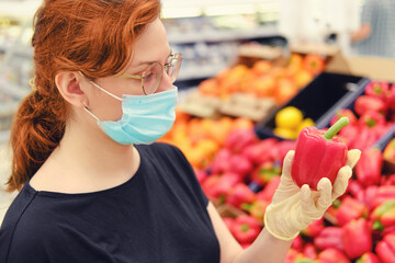 A young woman in a medical mask between rows of vegetables in a supermarket. Girl chooses bell pepper in the store. Flu virus quarantine in public places