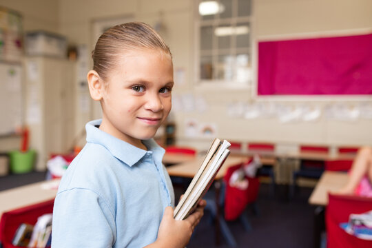 Girl school student in a classroom holding a set of books looking at camera