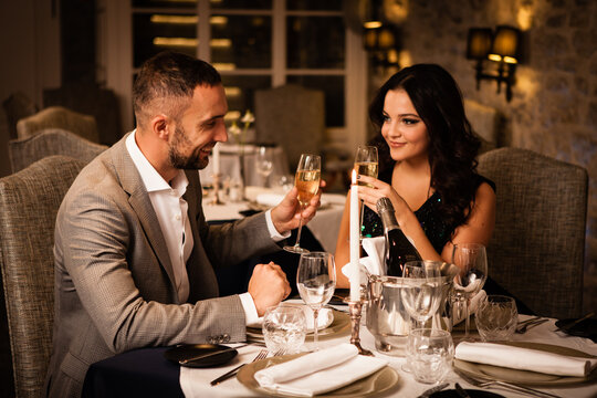 Romance And Dating Concept. Young Couple Man And Woman With Champagne Glasses Celebrating And Toasting In Restaurant. Two People Male And Female In Elegant Evening Suit And Dress On Romantic Dinner